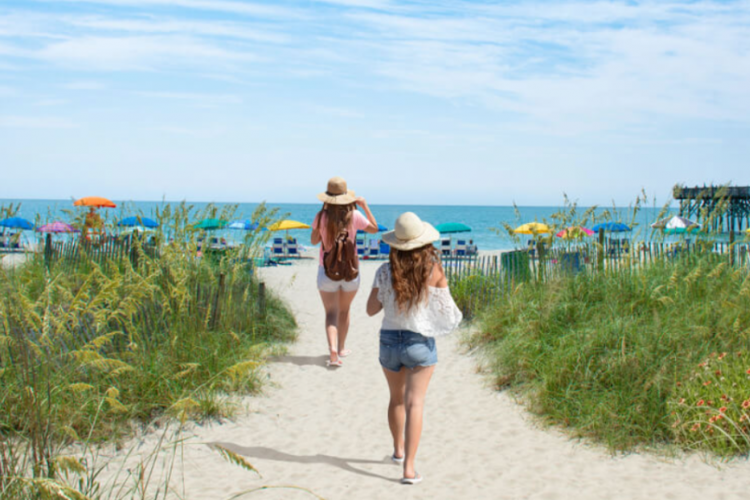 two girls walking onto the beach, in the background we see colorful umbrellas along the shoreline